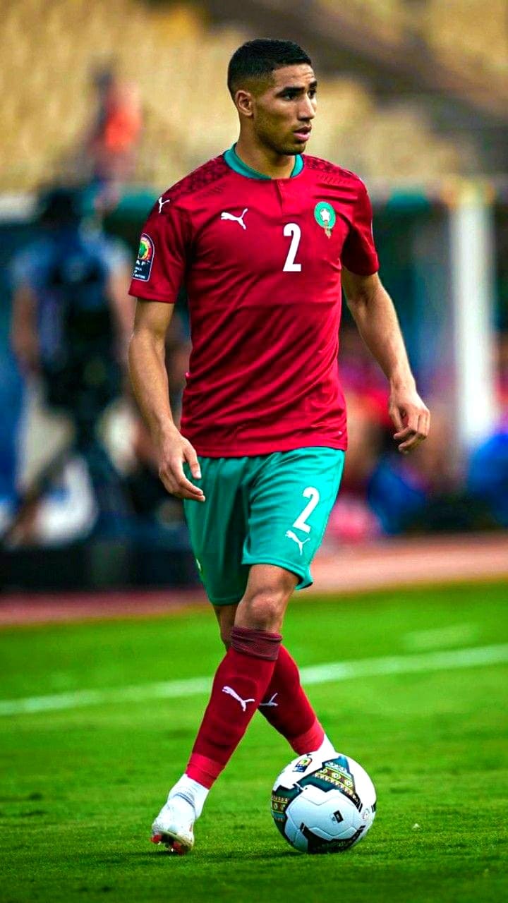 A soccer player in a red and green Morocco national team uniform with the number 2 dribbles a ball on the field during an IPTV Latino-broadcast match.
